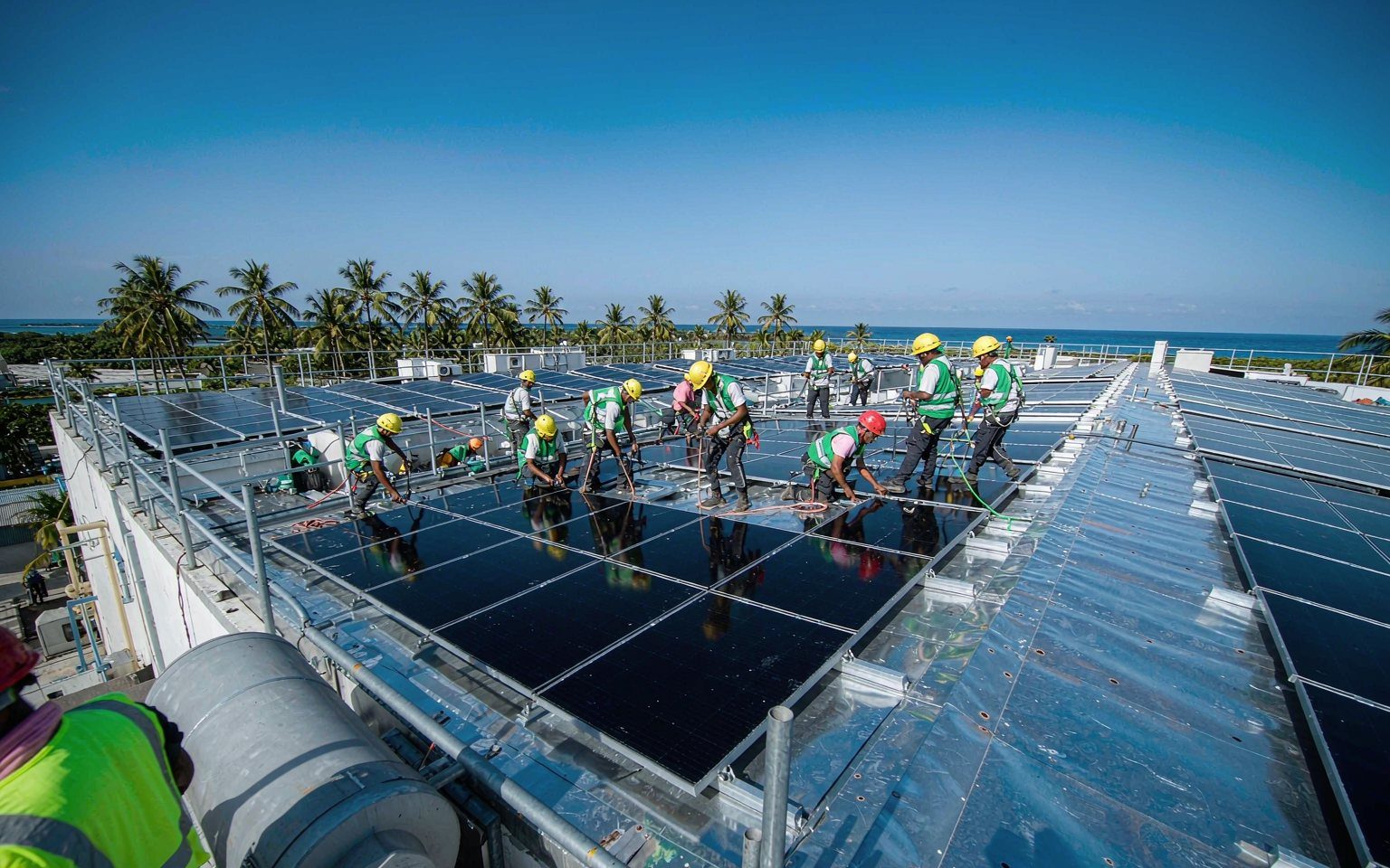 Technicians installing solar panels on a large commercial rooftop in a tropical Pacific island setting